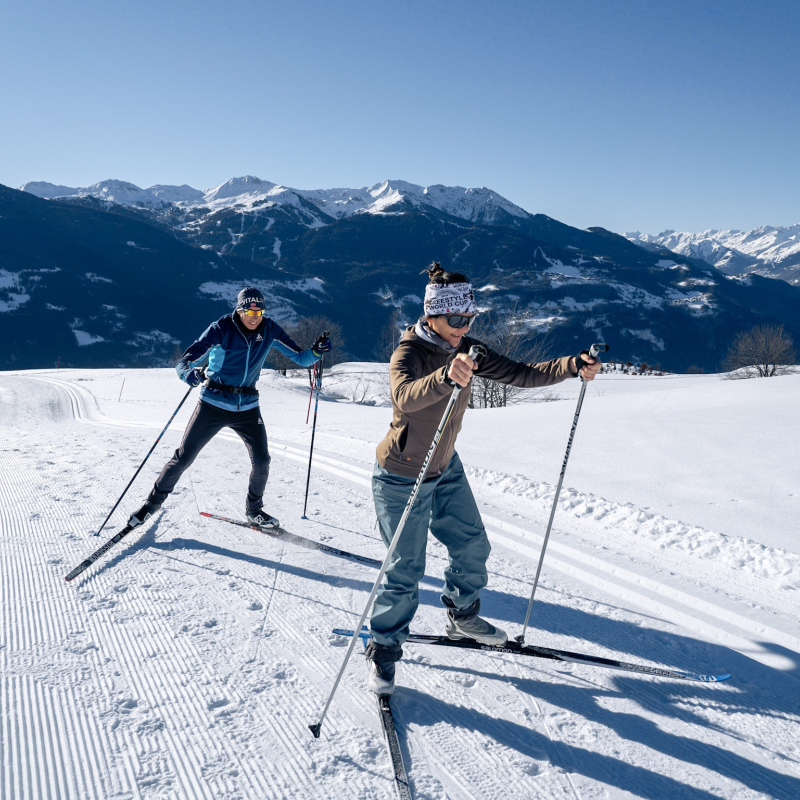 Couple de fondeurs sur une piste enneigée avec vue dégagée sur une chaine de montagne.